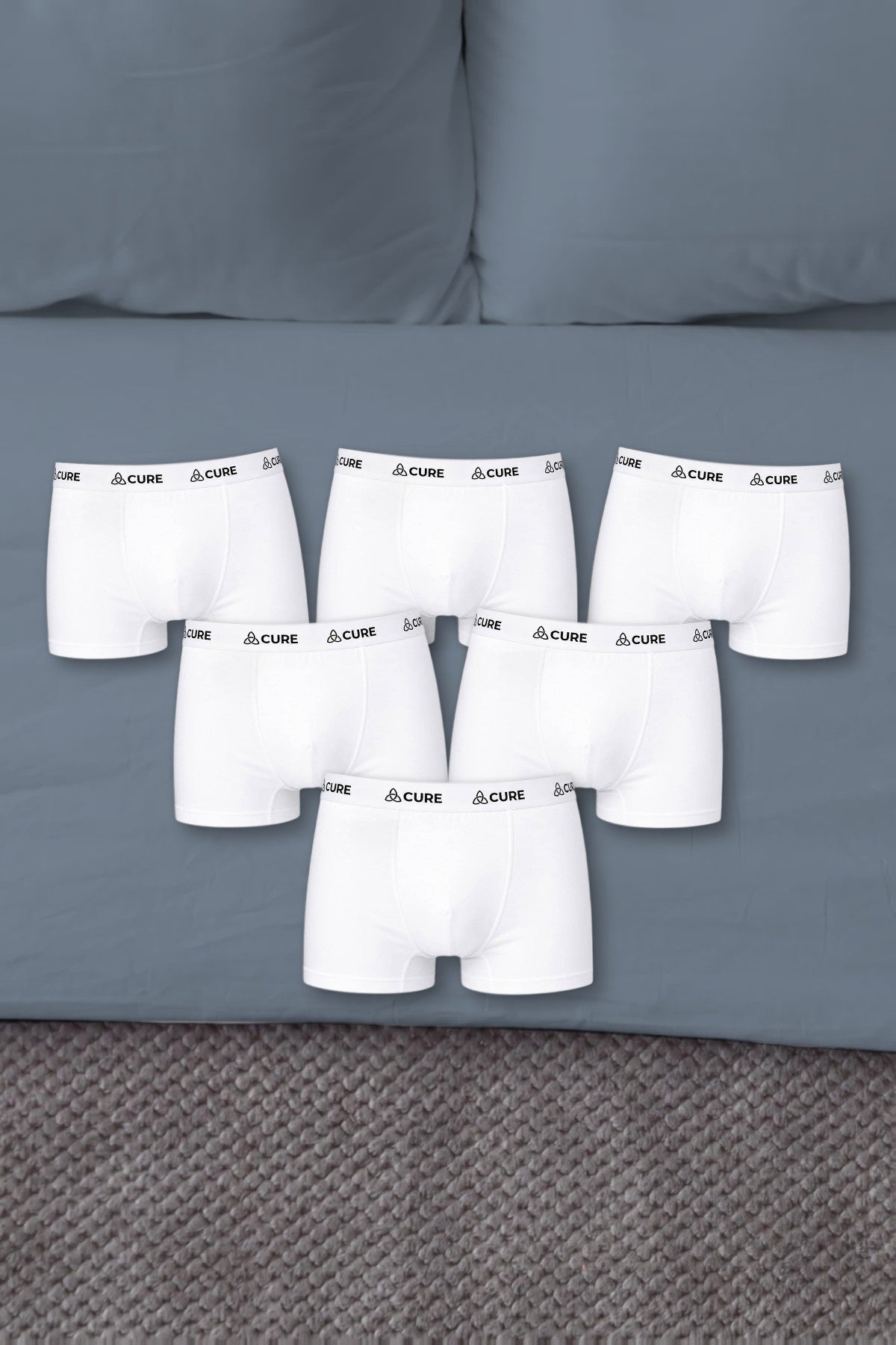 Sechs weiße ComfortCure Boxershorts auf einem Bett mit blaugrauer Bettwäsche