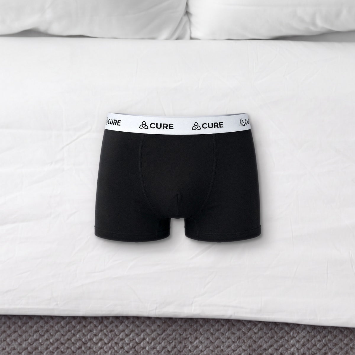 ComfortCure Herren-Boxershorts mit saugfähiger Innenschicht in Schwarz, liegen auf einem Bett mit weißen Kissen