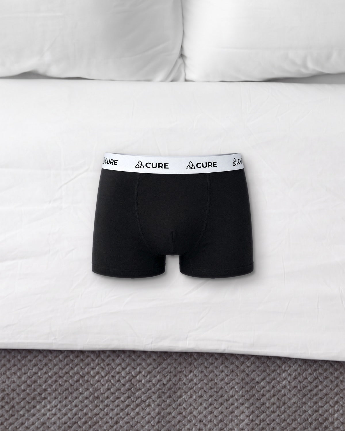 ComfortCure Herren-Boxershorts mit saugfähiger Innenschicht in Schwarz, liegen auf einem Bett mit weißen Kissen