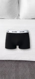 ComfortCure Herren-Boxershorts mit saugfähiger Innenschicht in Schwarz, liegen auf einem Bett mit weißen Kissen