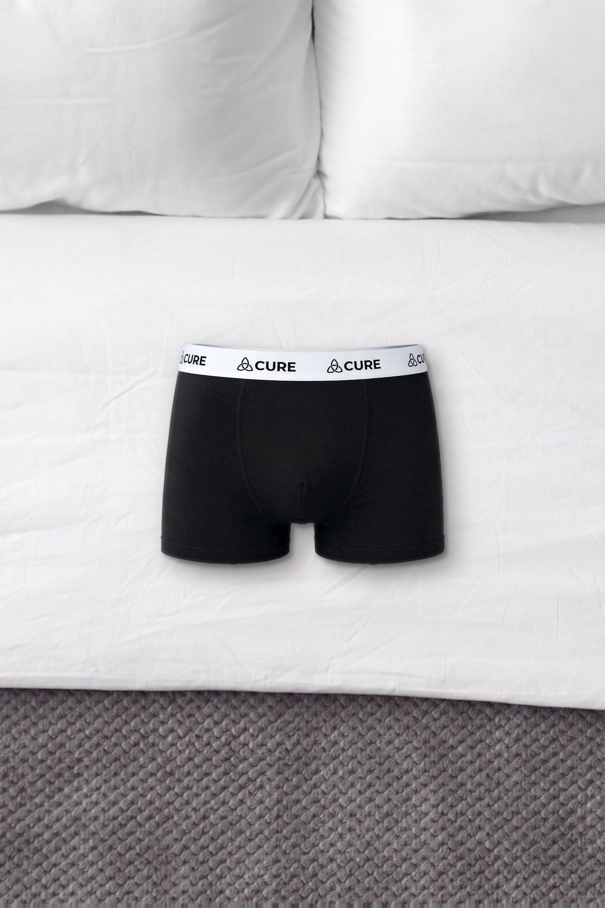 ComfortCure Herren-Boxershorts mit saugfähiger Innenschicht in Schwarz, liegen auf einem Bett mit weißen Kissen