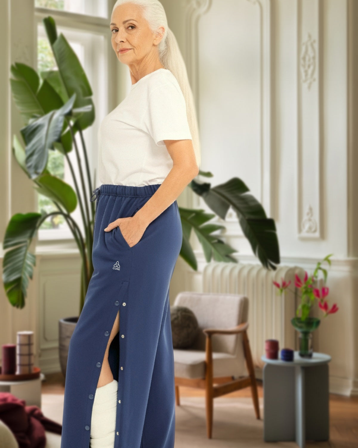 ComfortCure Adaptive Orthopädie-Pflegehose mit Druckknöpfen, Navy, Seitenansicht im Altbau-Wohnzimmer, seitlich geöffnet über einem bandagierten Bein