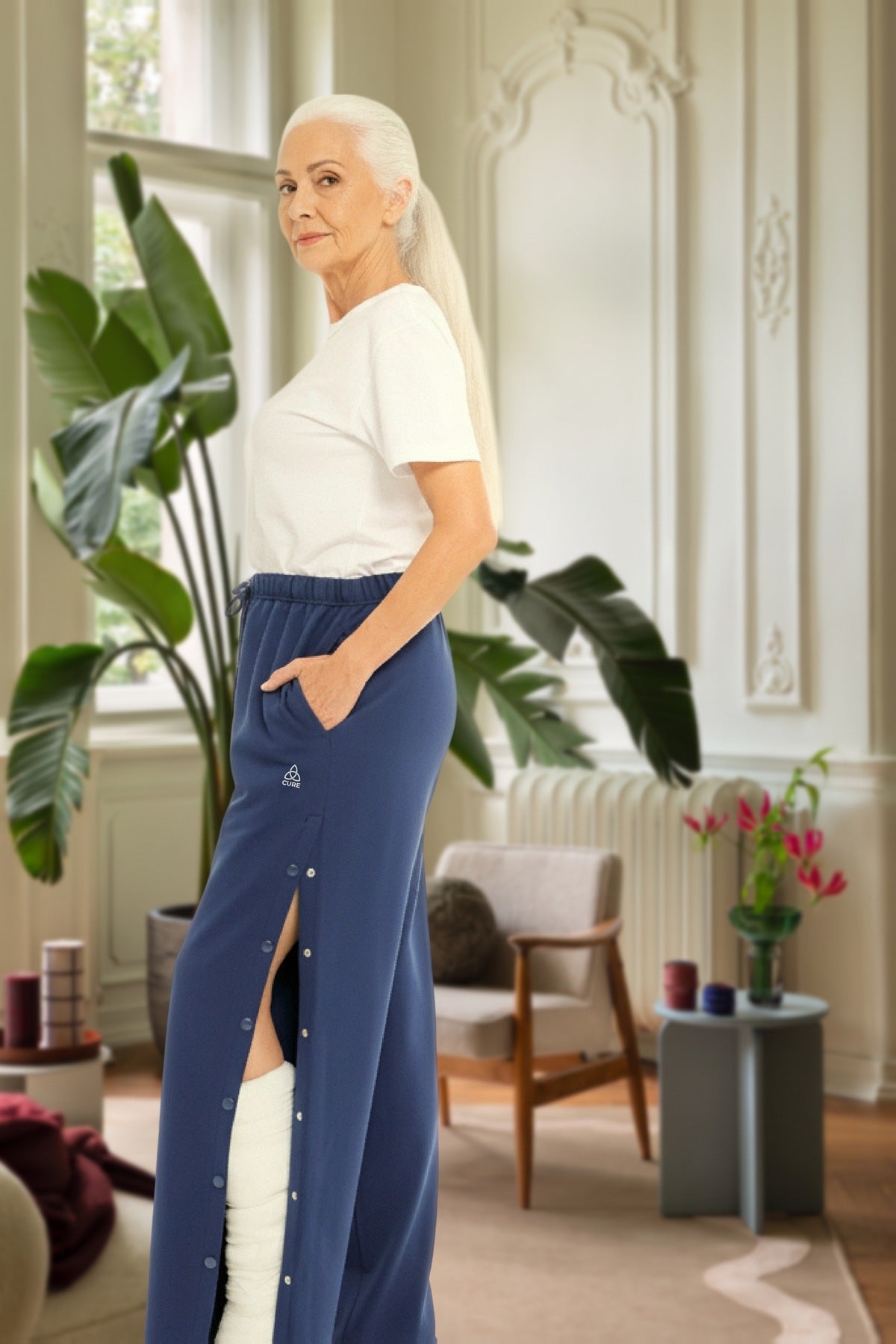 ComfortCure Adaptive Orthopädie-Pflegehose mit Druckknöpfen, Navy, Seitenansicht im Altbau-Wohnzimmer, seitlich geöffnet über einem bandagierten Bein