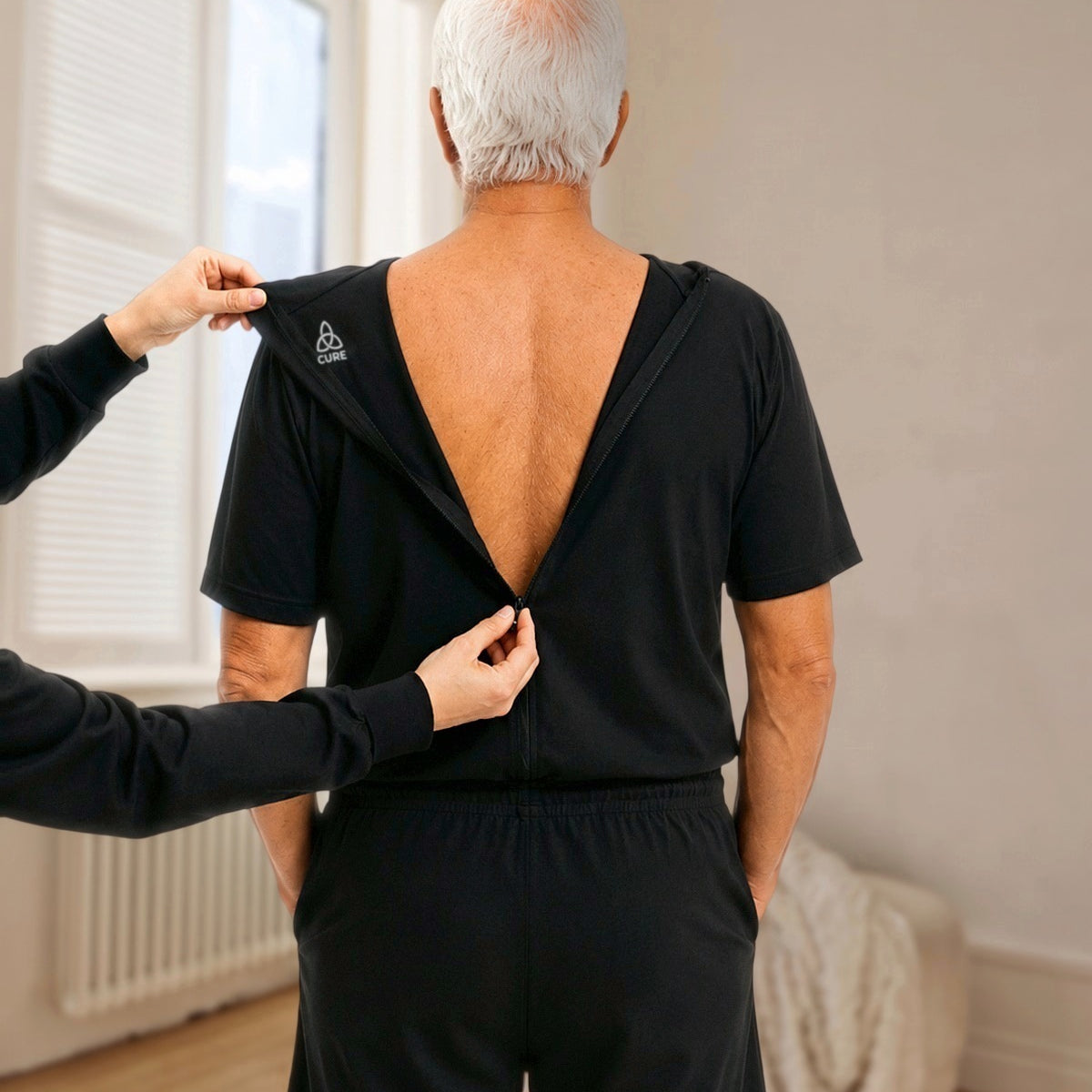 ComfortCure Adaptiver Kurzarm-Pflegeoverall für die Alzheimer- und Demenzpflege, Schwarz, Rückenansicht im hellen Altbauzimmer, eine Pflegekraft öffnet den Rückenreißverschluss