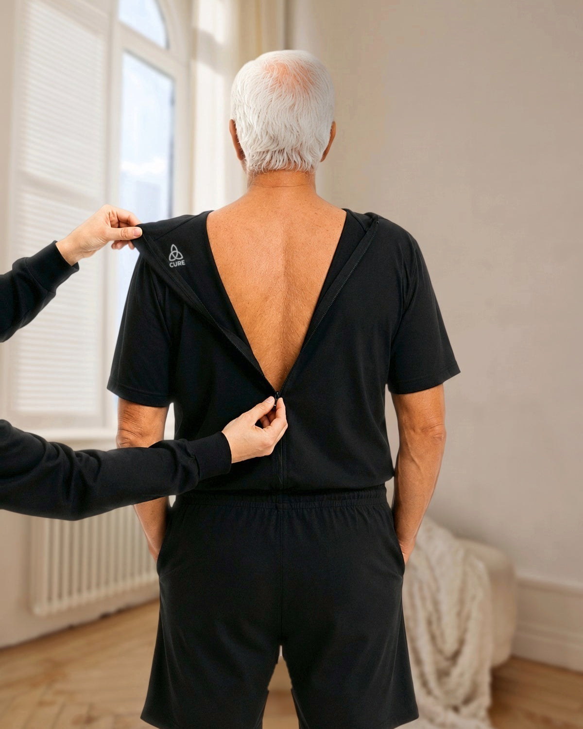 ComfortCure Adaptiver Kurzarm-Pflegeoverall für die Alzheimer- und Demenzpflege, Schwarz, Rückenansicht im hellen Altbauzimmer, eine Pflegekraft öffnet den Rückenreißverschluss