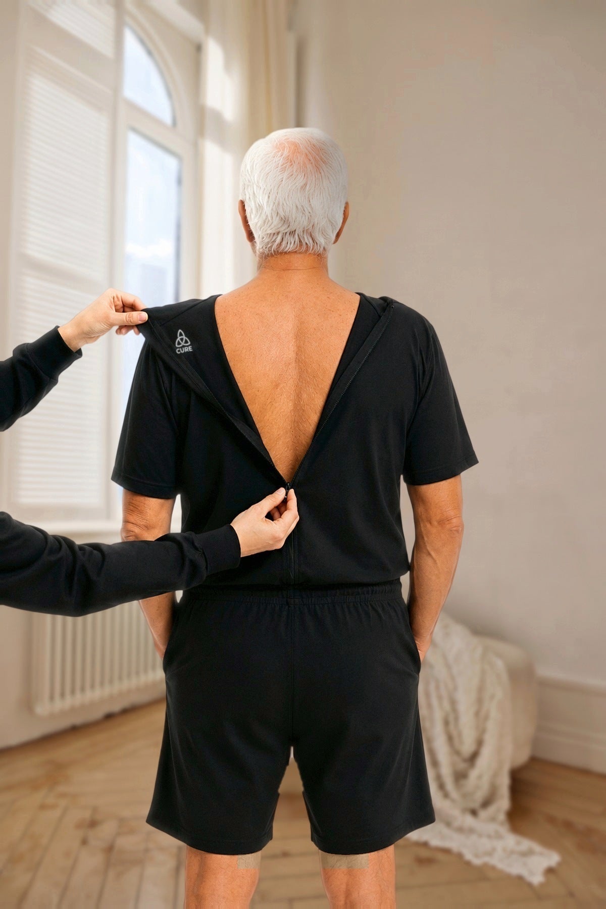 ComfortCure Adaptiver Kurzarm-Pflegeoverall für die Alzheimer- und Demenzpflege, Schwarz, Rückenansicht im hellen Altbauzimmer, eine Pflegekraft öffnet den Rückenreißverschluss