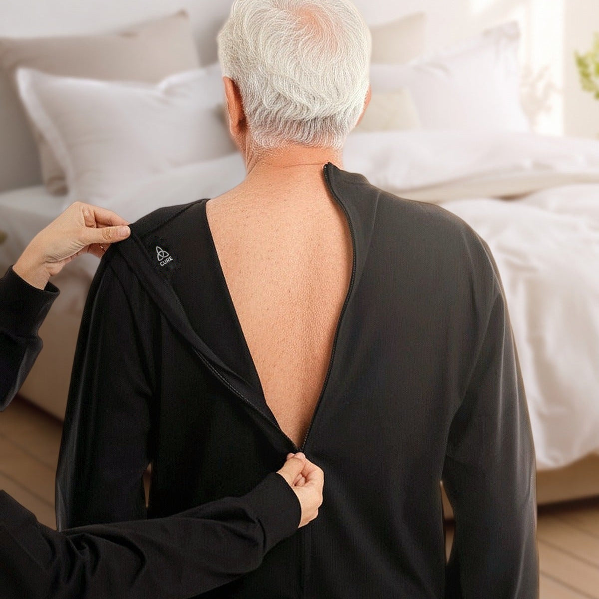 ComfortCure Pflegeoverall Langarm in Schwarz, Rückansicht im Schlafzimmer, eine Helferin öffnet den Reißverschluss am Rücken