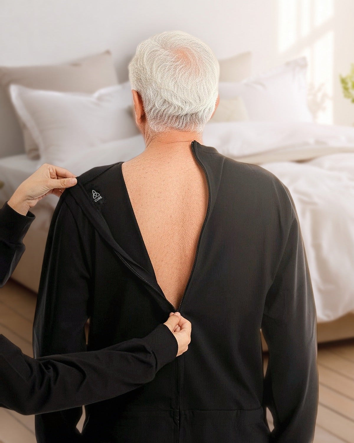 ComfortCure Pflegeoverall Langarm in Schwarz, Rückansicht im Schlafzimmer, eine Helferin öffnet den Reißverschluss am Rücken