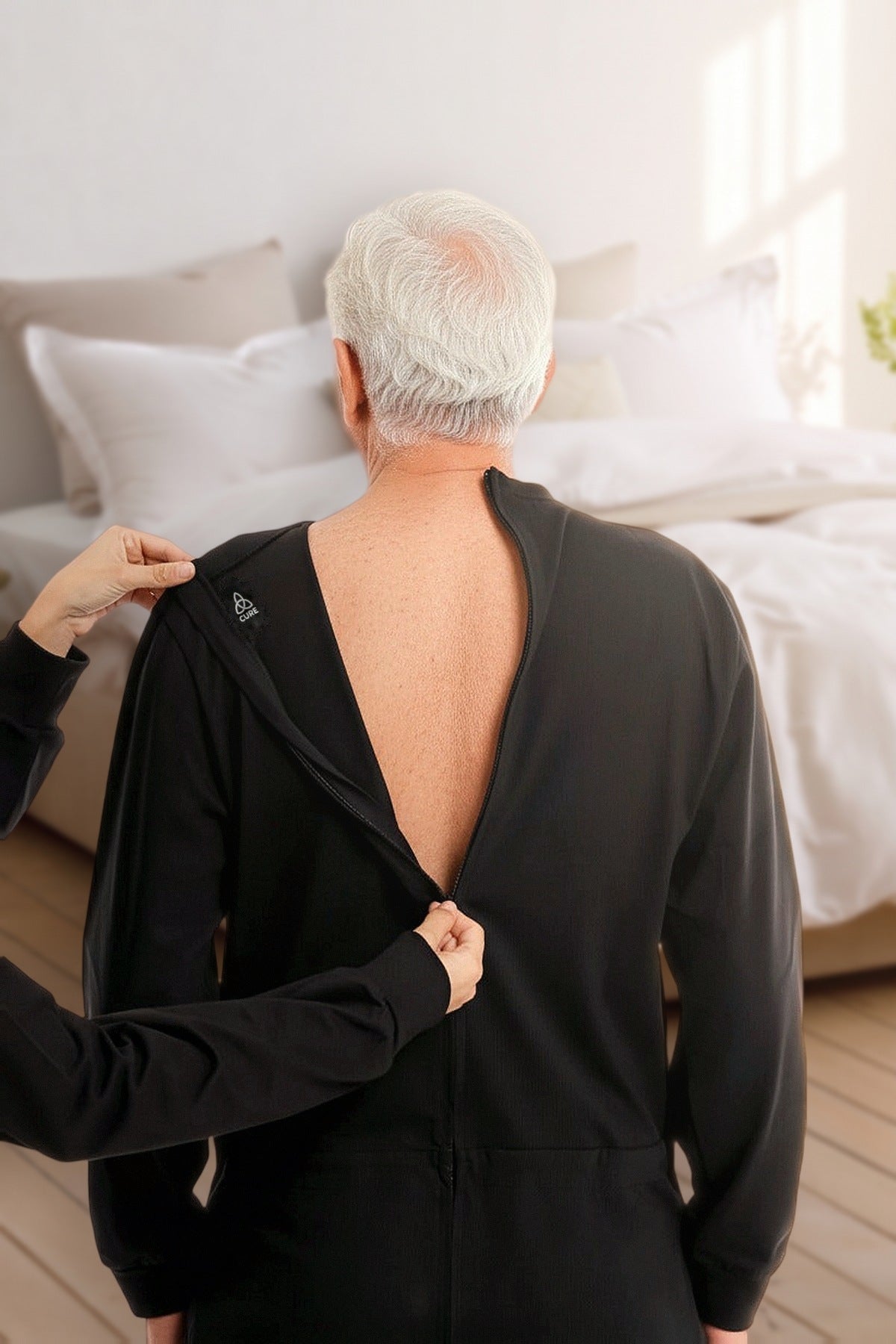 ComfortCure Pflegeoverall Langarm in Schwarz, Rückansicht im Schlafzimmer, eine Helferin öffnet den Reißverschluss am Rücken